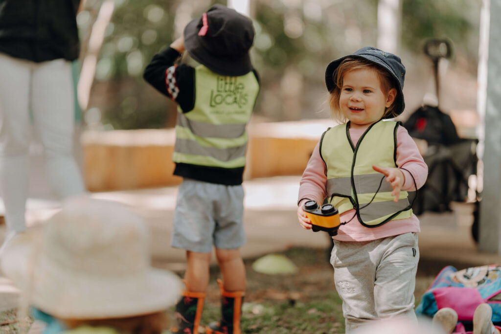 A child wearing a Little Locals’ Early Learning high-vis vest holds binoculars in preparation for a Bush Kindy excursion with the long daycare centre.