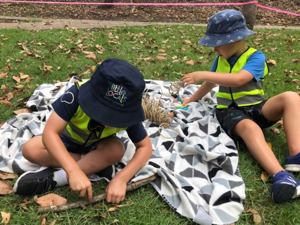 Two children wearing Little Locals’ Early Learning high-vis vests sit on a picnic blanket creating crafts with string during a Bush Kindy excursion at the long daycare centre.