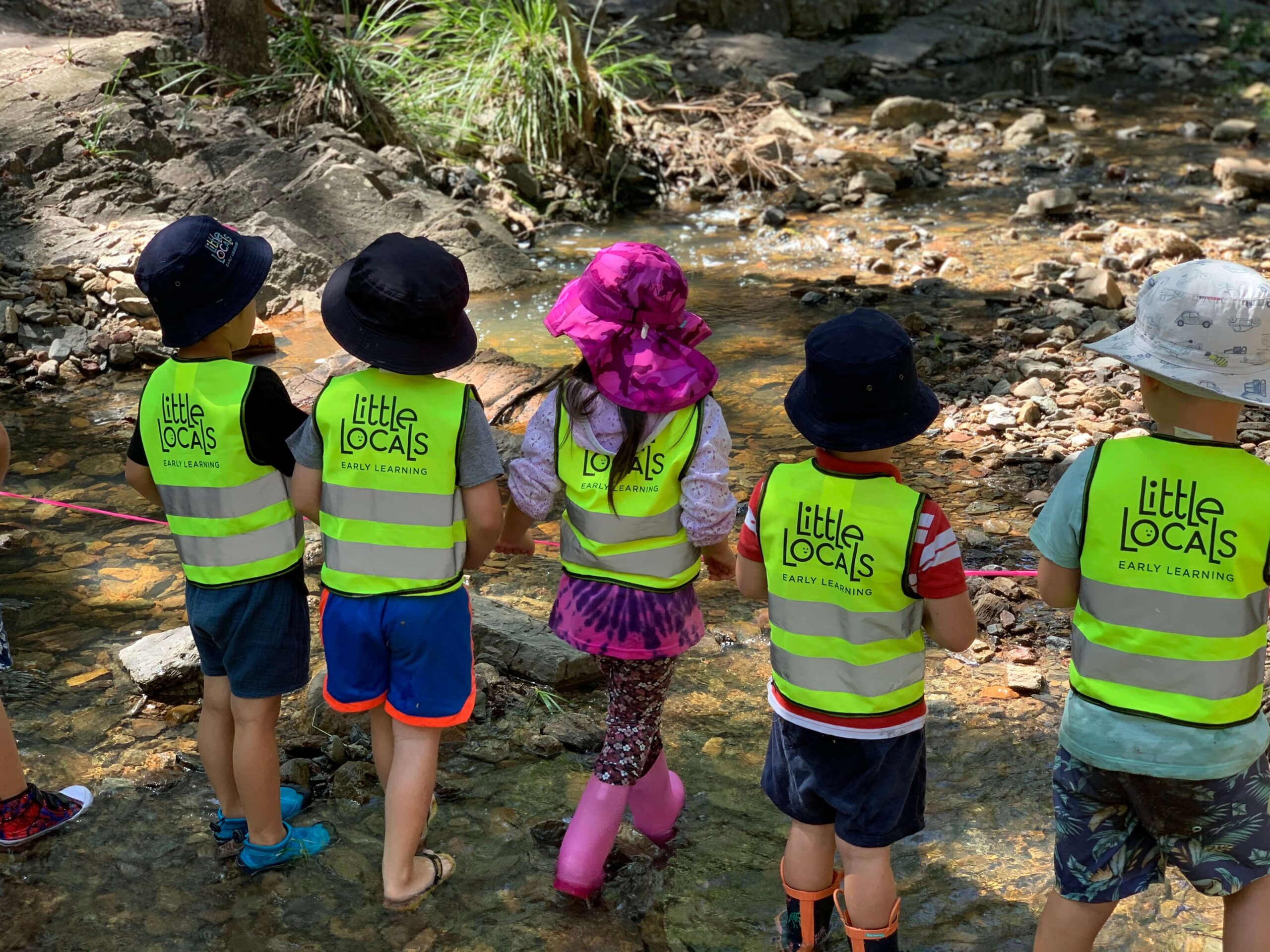 A group of children wearing Little Locals’ Early Learning high-vis vests stand holding a piece of string together just before a creek during a Bush Kindy excursion at the long daycare centre.