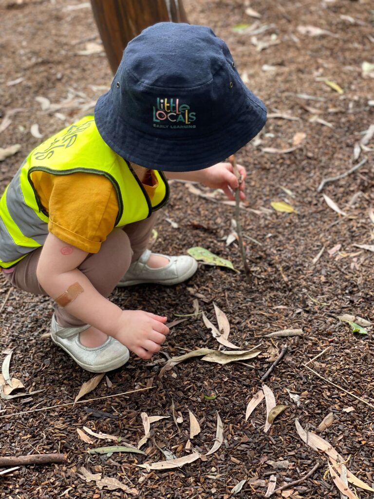 A child wearing a Little Locals’ Early Learning high-vis vest plays with sticks and leaves during a Bush Kindy excursion at the long daycare centre.