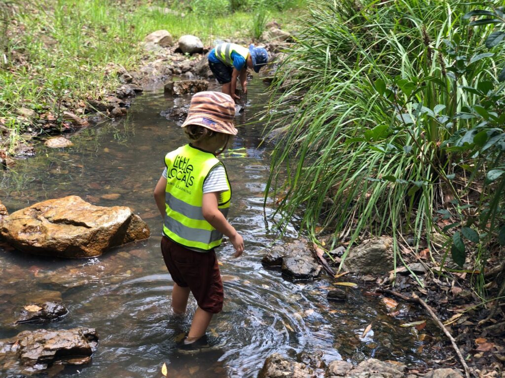 A child wearing a Little Locals’ Early Learning high-vis vest stands in a creek during a Bush Kindy excursion at the long daycare centre.