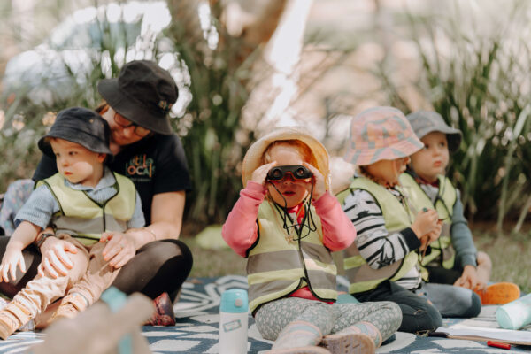A group of children from Little Locals Early Learning Centre sits on a picnic rug during an outdoor excursion to learn more about sustainability and the environment.